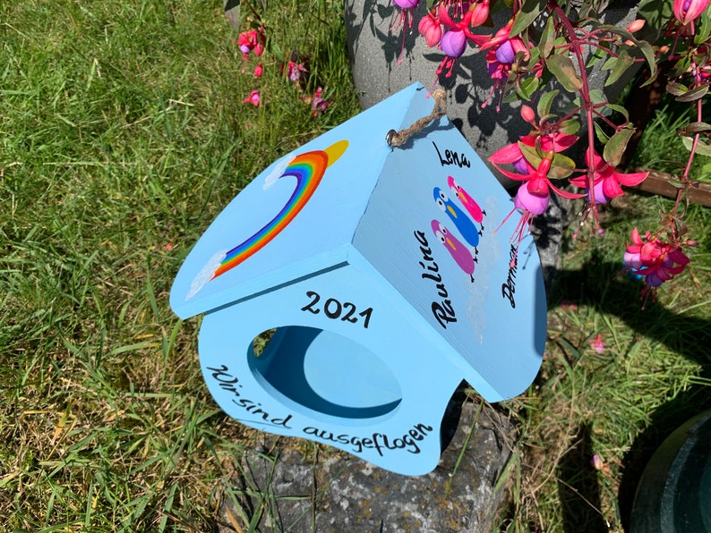 May include: A light blue wooden birdhouse featuring a painted rainbow, the year 2021, and the German text "Wir sind ausgeflogen". The roof displays three painted bird figures with the names Lena, Paulina, and Damian written in black script.