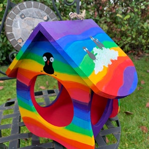 May include: A wooden birdhouse painted with vibrant rainbow stripes in red, orange, yellow, green, blue, and purple. The roof features hand-painted black bird figures and a white cloud. It has a large circular opening and a jute hanging loop.