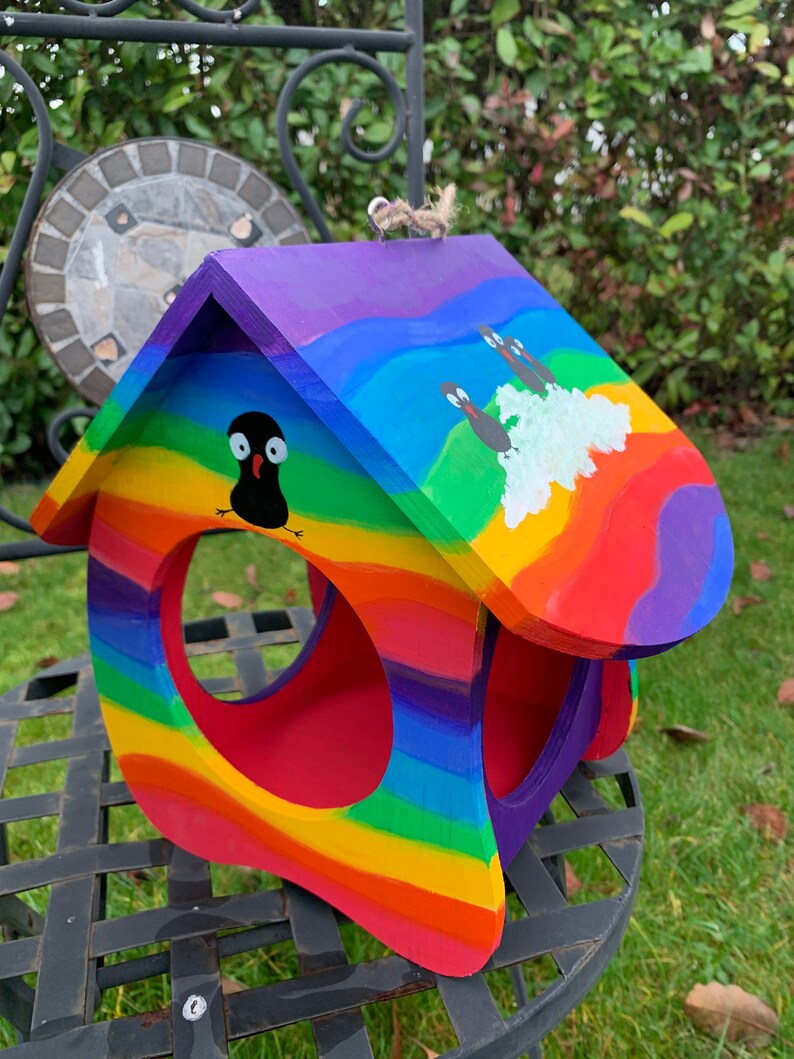 May include: A wooden birdhouse painted with vibrant rainbow stripes in red, orange, yellow, green, blue, and purple. The roof features hand-painted black bird figures and a white cloud. It has a large circular opening and a jute hanging loop.