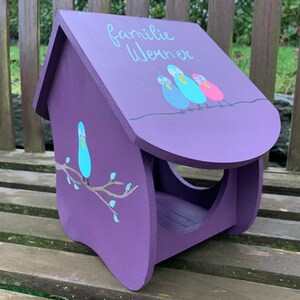 May include: A purple wooden bird feeder with a slanted roof featuring the painted text "familie Werner" and three colorful birds perched on a line. The side panel displays a single blue bird sitting on a brown branch with small light blue leaves.