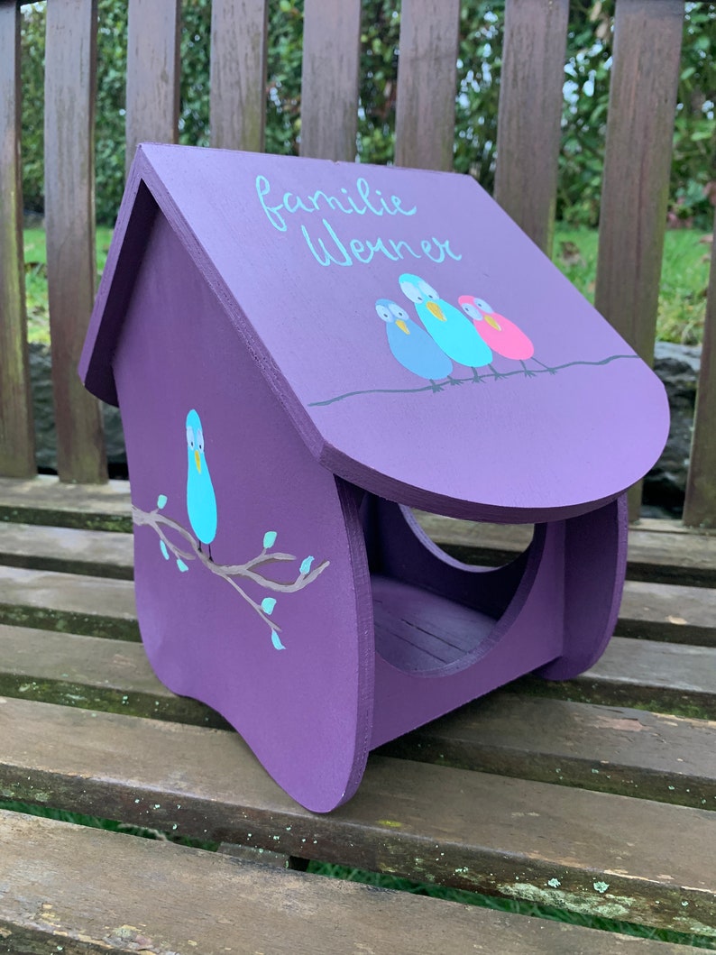 May include: A purple wooden bird feeder with a slanted roof featuring the painted text "familie Werner" and three colorful birds perched on a line. The side panel displays a single blue bird sitting on a brown branch with small light blue leaves.