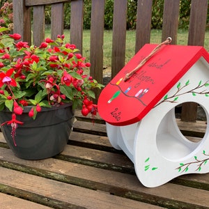 May include: A white wooden birdhouse with a red roof featuring a hand-painted branch with small birds and the German text Wir sind ausgeflogen 2020. The birdhouse has a large circular opening on the front and is decorated with painted green leaves.