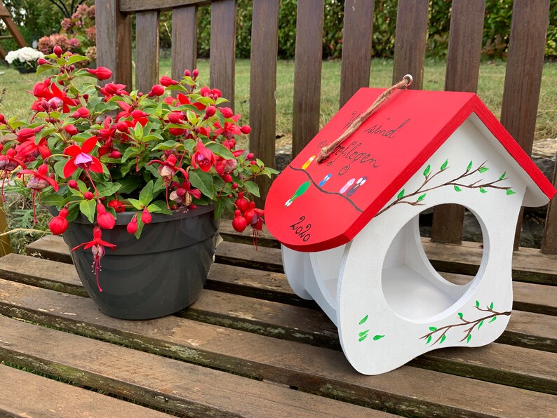 May include: A white wooden birdhouse with a red roof featuring a hand-painted branch with small birds and the German text Wir sind ausgeflogen 2020. The birdhouse has a large circular opening on the front and is decorated with painted green leaves.