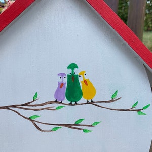 May include: A white wooden birdhouse with a red roof featuring a hand-painted illustration of three cartoon birds in purple, green, and yellow perched on a brown branch with small green leaves. The birds have large white eyes and orange beaks.
