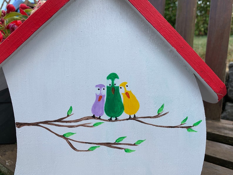 May include: A white wooden birdhouse with a red roof featuring a hand-painted illustration of three cartoon birds in purple, green, and yellow perched on a brown branch with small green leaves. The birds have large white eyes and orange beaks.