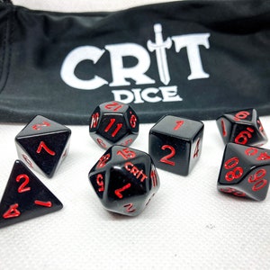 以下が含まれることがあります： 赤い数字の黒いサイコロのセット。サイコロは、d4、d6、d8、d10、d12、d20など、さまざまな形をしています。サイコロは白い表面の上にあります。サイコロはCrit Dice製です。