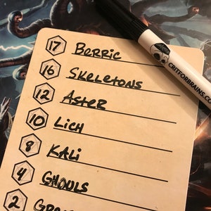 May include: A brown card with a list of names and numbers written in black marker. The numbers are in a hexagonal shape and the names are written next to them. The card is on a table with a black marker pen with a skull logo and the text "CRITFORBRAINS.COM" on it.