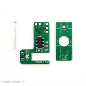 Peut inclure: Circuit imprimé vert avec un interrupteur d'alimentation, un tube en plastique transparent et un gabarit de perçage vert avec des marques pour des trous de 3 mm et 9 mm. Le circuit imprimé porte l'inscription "SMS2 REGION セガ".