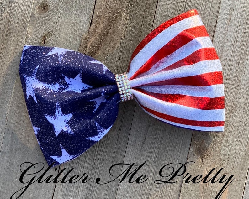 May include: A patriotic hair bow featuring a design inspired by the American flag. The bow has a navy blue section with white stars and a red and white striped section. A sparkling rhinestone embellishment adds a touch of glamour.