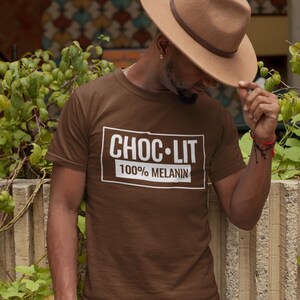 May include: A brown t-shirt with a white box that says "CHOC-LIT 100% MELANIN" on it. The person wearing the shirt is wearing a brown fedora.