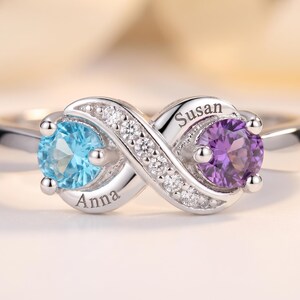 May include: A silver infinity ring featuring a round light blue gemstone on one side and a round purple gemstone on the other. The band is accented with small clear stones and includes the engraved names Anna and Susan on the metal surface.