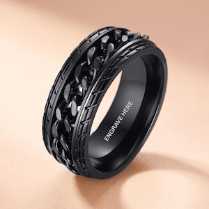 May include: A black ring featuring a central rotating chain-link band flanked by two grooved edges. The inner surface of the ring is smooth and displays the text ENGRAVE HERE in a clean, sans-serif font, indicating a space for custom personalization.