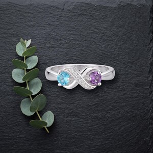 May include: A silver-toned ring featuring an infinity-shaped design set with a round light blue gemstone on one side and a round purple gemstone on the other, accented with small clear stones along the center band.