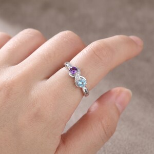 May include: A silver-toned infinity-shaped ring featuring a round purple gemstone and a round light blue gemstone. The band is accented with small clear stones and includes the engraved text Name on the side of the metal band.