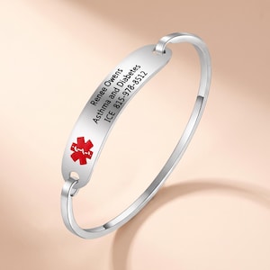 May include: A silver-toned metal medical alert bangle bracelet featuring a flat, curved identification plate. The plate displays a red medical star of life symbol and engraved text: Renee Owens, Asthma and Diabetes, ICE 815-978-8512.