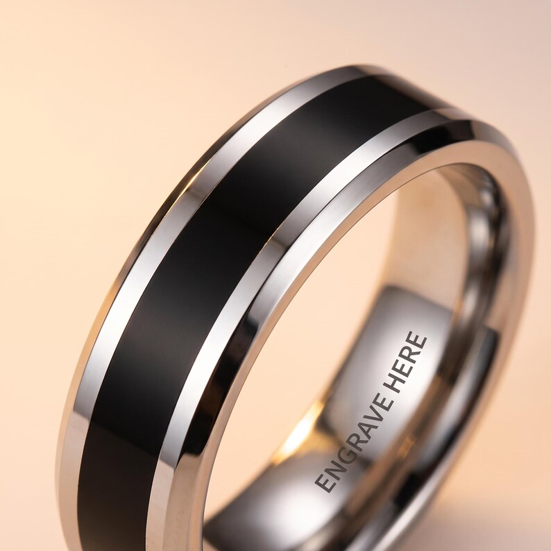 Może przedstawiać: Srebrzysty metalowy pierścień z szerokim, matowym czarnym pasem środkowym otoczonym polerowanymi srebrnymi krawędziami. Wewnętrzna powierzchnia pierścienia jest gładka i metaliczna, wyraźnie prezentując napis ENGRAVE HERE czcionką bezszeryfową.