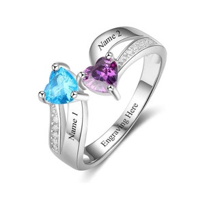 May include: A silver ring with two heart-shaped gemstones, one blue and one purple. The ring has two engraved names, "Name 1" and "Name 2", and the words "Engraving Here" on the band.