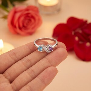 May include: A silver-toned ring featuring an infinity-shaped design set with a round light blue gemstone on one side and a round purple gemstone on the other, accented with small clear stones along the center of the infinity symbol band.
