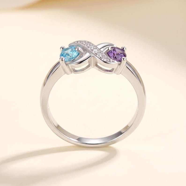 May include: A silver-toned ring featuring an infinity symbol design. The band is accented with small clear stones along the top curve. One side of the infinity loop holds a round light blue stone, while the other side holds a round purple stone.