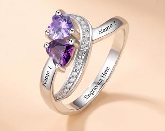 May include: A silver-toned ring featuring two heart-shaped purple gemstones, one light and one dark, set alongside a band encrusted with small clear stones. The metal band includes the engraved text Name 1 and Engraving Here.