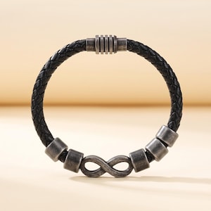 May include: A black braided leather bracelet featuring a metallic infinity symbol charm, four cylindrical silver-toned beads, and a ribbed barrel clasp. The bracelet is constructed from a single woven leather cord with a dark, weathered finish.