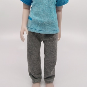 May include: A doll with blonde hair wearing a blue and turquoise t-shirt and gray pants.