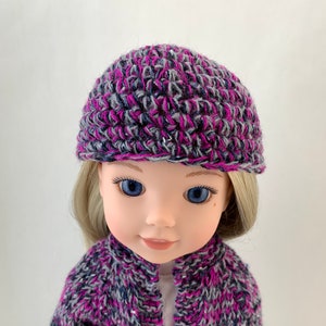 Op de afbeelding: Een pop die een gebreide cardigan draagt in paars, grijs en zwart met een bijpassende paarse, grijze en zwarte gehaakte muts.