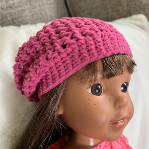May include: A pink crocheted beanie hat with a textured design. The hat is worn on a doll with brown hair and brown eyes.
