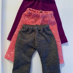 May include: Three pairs of pants in different colors and patterns. The bottom pair is black and white striped, the middle pair is pink with a white pattern, and the top pair is purple.