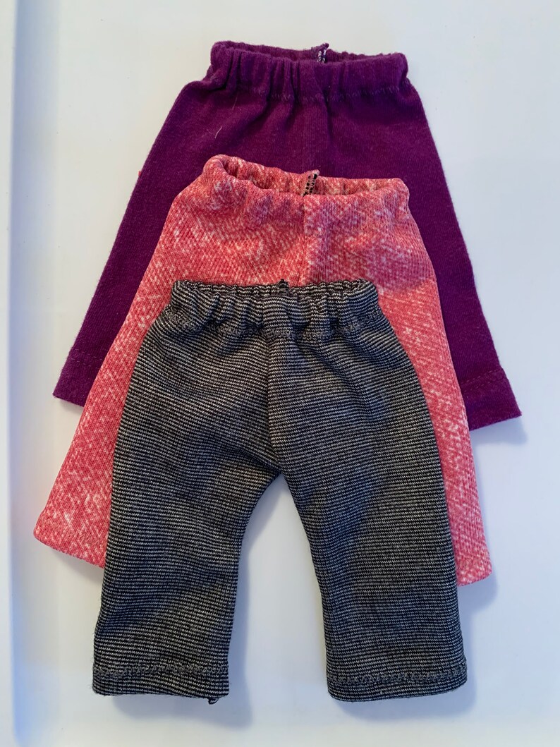 May include: Three pairs of pants in different colors and patterns. The bottom pair is black and white striped, the middle pair is pink with a white pattern, and the top pair is purple.