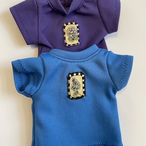 May include: Two short-sleeved shirts with embroidered flower patches. The top shirt is purple and the bottom shirt is blue.