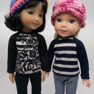 May include: Two dolls wearing striped shirts and crocheted hats. One doll has black hair and is wearing a black and white floral shirt and black pants. The other doll has blonde hair and is wearing a blue and white striped shirt and blue jeans.