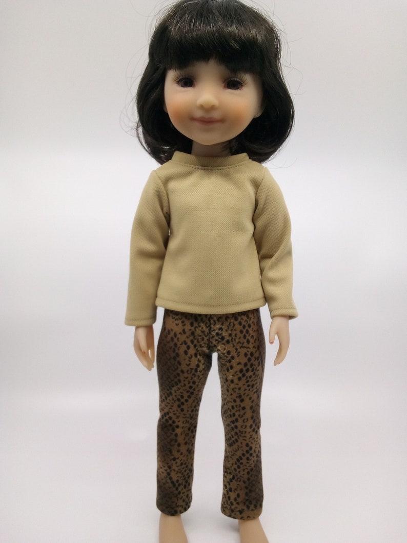 May include: A doll wearing a beige long-sleeved shirt and brown and black patterned pants.