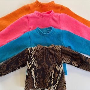 Puede incluir: Cuatro camisas de manga larga apiladas. La camisa de arriba es naranja, la segunda es rosa, la tercera es azul y la última tiene un estampado de piel de serpiente marrón y blanco.