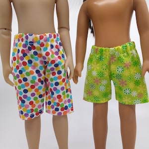 May include: Two dolls wearing different patterned shorts. One doll is wearing colorful polka dot shorts and the other is wearing green shorts with a floral pattern.