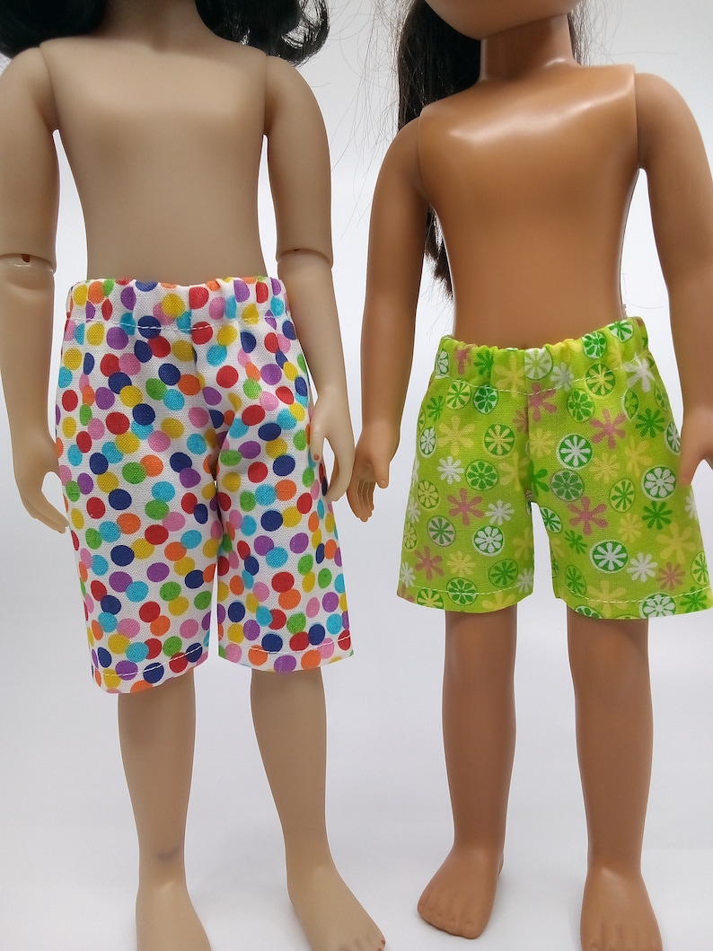 May include: Two dolls wearing different patterned shorts. One doll is wearing colorful polka dot shorts and the other is wearing green shorts with a floral pattern.