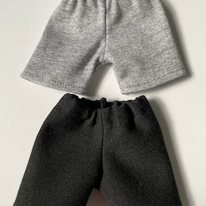 Peut inclure: Deux paires de shorts pour poupées. Une paire est gris chiné, l'autre est noire. Les deux paires ont une taille élastique et un design simple et classique. Les shorts sont faits d'un tissu doux et texturé.