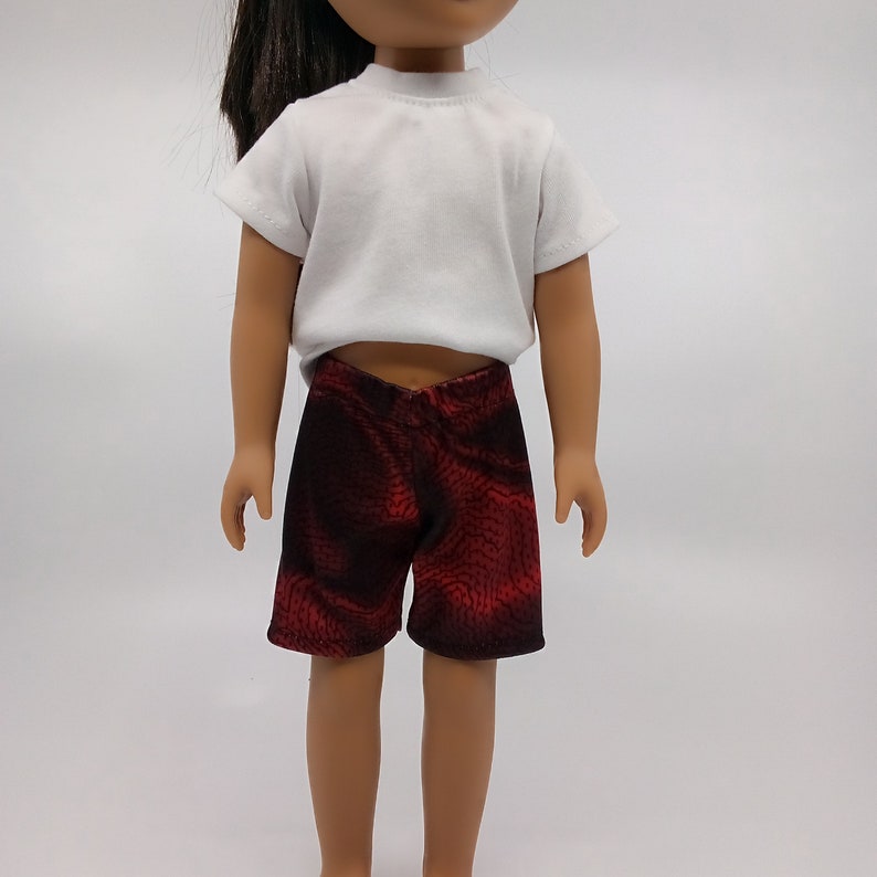 May include: A doll wearing a white short-sleeved shirt and red and black patterned shorts.