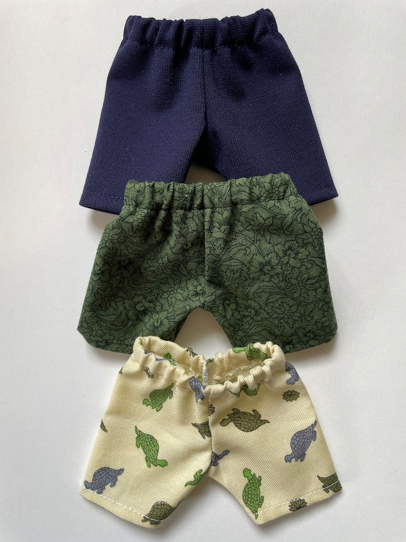 May include: Three pairs of shorts in different colors and patterns. The top pair is navy blue, the middle pair is green with a floral pattern, and the bottom pair is cream with a green turtle pattern.