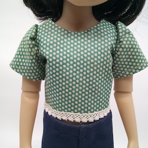 May include: A doll wearing a green polka dot top with a white lace trim and dark blue pants. The top has short sleeves and a rounded neckline. The doll's arms are visible.