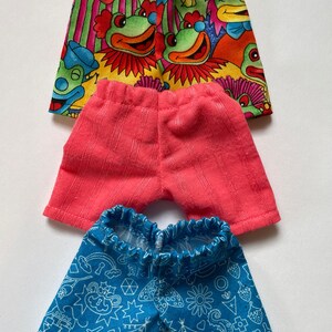May include: Three pairs of doll shorts. The first pair is multi-colored with a clown and frog pattern. The second pair is a solid pink. The third pair is blue with a white geometric pattern.