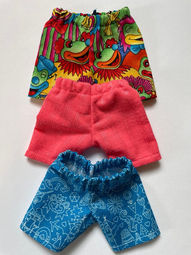 May include: Three pairs of doll shorts. The first pair is multi-colored with a clown and frog pattern. The second pair is a solid pink. The third pair is blue with a white geometric pattern.