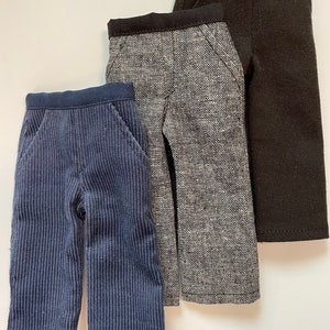 Può includere: Tre paia di pantaloni in tessuti e colori diversi. I pantaloni a sinistra sono di velluto a coste blu, quelli al centro sono di tweed grigio e quelli a destra sono neri.