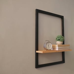 Può includere: Uno scaffale in metallo nero con un ripiano in legno, un piccolo orologio, una pianta in vaso e tre libri. Lo scaffale è montato su una parete di colore grigio chiaro.
