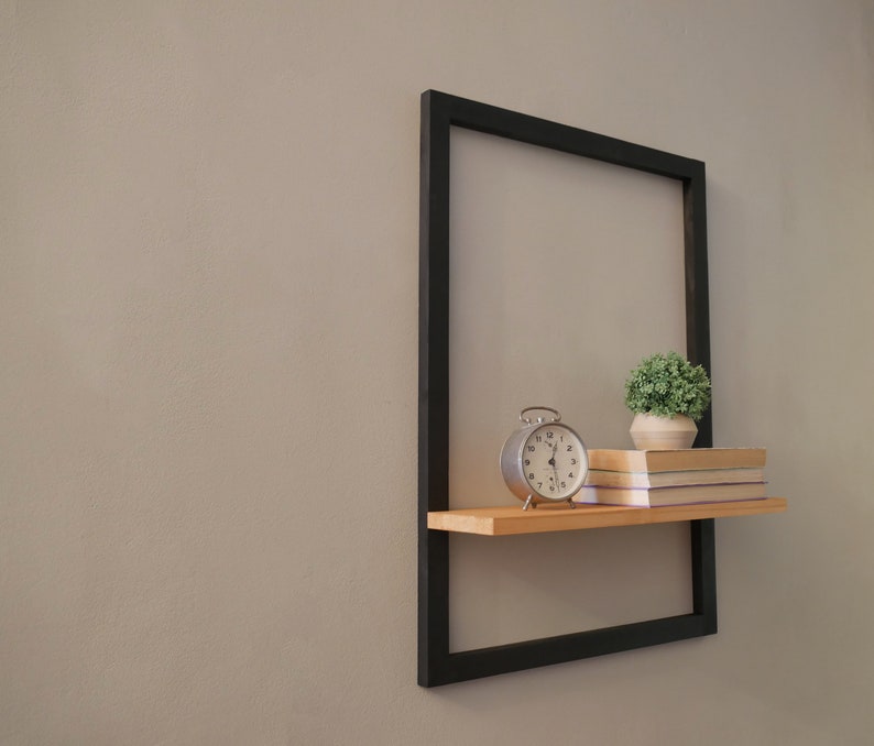 Puede incluir: Una estanter&iacute;a de metal negro con una repisa de madera, un peque&ntilde;o reloj, una planta en maceta y tres libros. La estanter&iacute;a est&aacute; montada en una pared de color gris claro.
