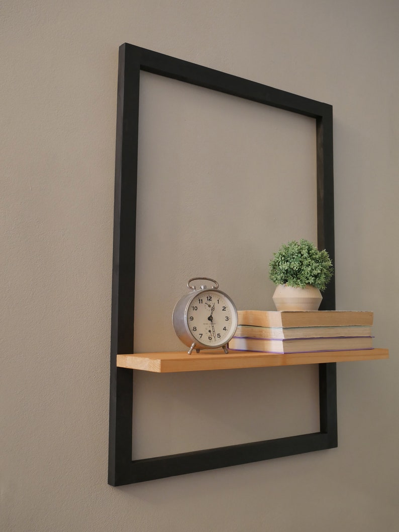 Puede incluir: Un marco de metal negro con un estante de madera. El estante tiene un reloj despertador plateado, una peque&ntilde;a planta en maceta y una pila de libros.