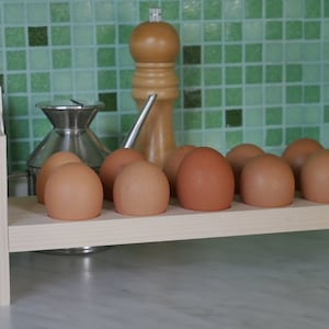 Könnte beinhalten: Ein Eierhalter aus Holz mit 10 braunen Eiern. Der Halter ist aus hellem Holz und hat ein schlichtes Design. Er steht auf einer weißen Arbeitsplatte.