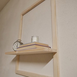 Puede incluir: Una estanter&iacute;a de madera con una vela blanca, una pila de libros y un reloj despertador plateado. La estanter&iacute;a est&aacute; unida a un marco de madera clara.