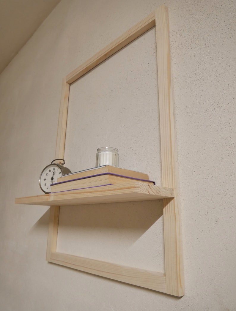 Puede incluir: Una estanter&iacute;a de madera con una vela blanca, una pila de libros y un reloj despertador plateado. La estanter&iacute;a est&aacute; unida a un marco de madera clara.
