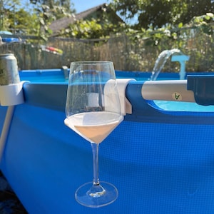 May include: A clear glass wine glass held by a white plastic clip-on holder attached to the blue metal frame of an above-ground swimming pool. The holder features a circular base and a curved arm that secures the stemware to the pool edge.